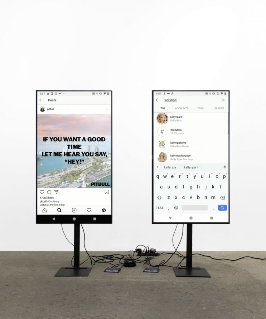 Fig. 5 Two vertical screens with their respective bases on the floor, connecting wires and two small apparatuses, providing a giant view of Pitbull and Kelly Ripa’s Instagram pages as they are seen on a phone.