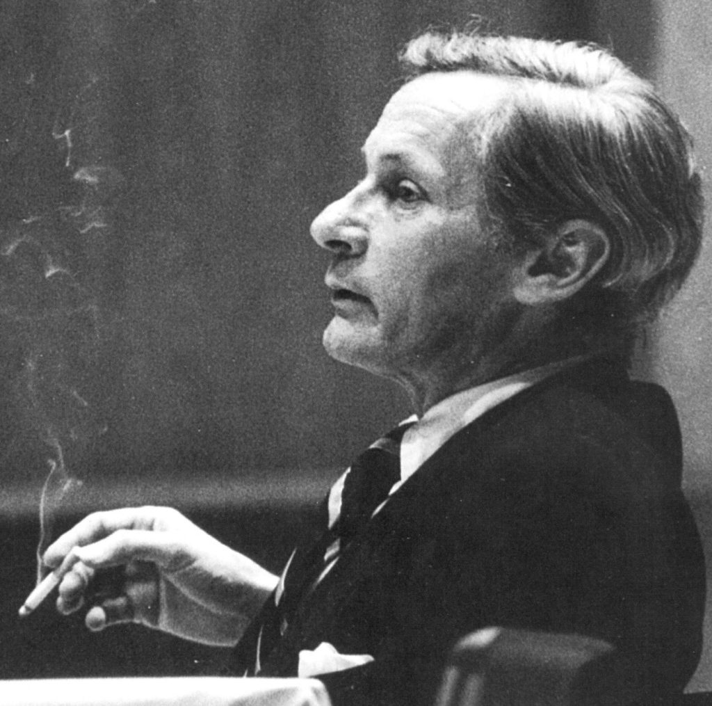 William Gaddis in profile, black and white. Smoking a cigarette in a suit, hair parted to the side. 